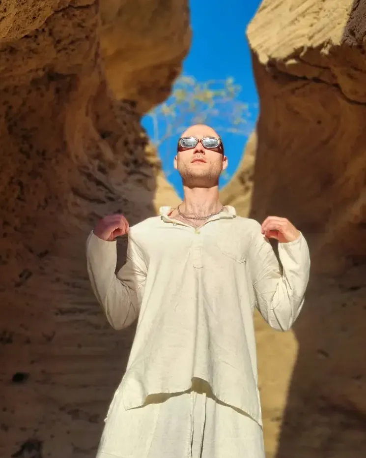 Kuba in white linen, sunglasses lowered, standing in a sandstone canyon